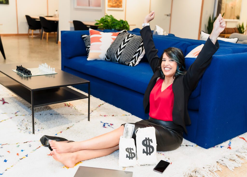 woman seated in the carpet and leaned on the soda, arms up, celebrating financial freedon