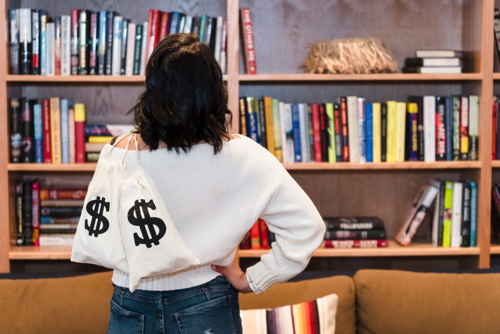 brand photo of a business Coach on the back looking at a library, holding 2 bags with money signs that represent her business

