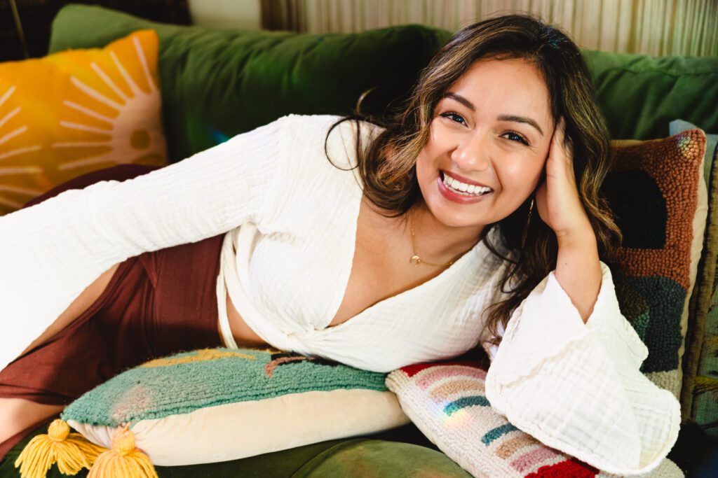 A woman lays down on a green sofa, hands on her head and a huge smile