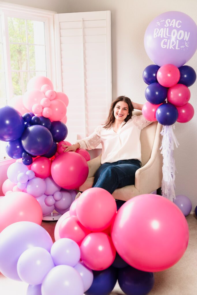 brand photo of a small business owner sitting in a chair among a lot of purple and pink balloons