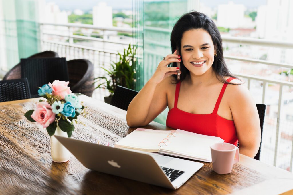 A woman sits in a wooden desk, on her phone, laptop, and agenda on the desk
