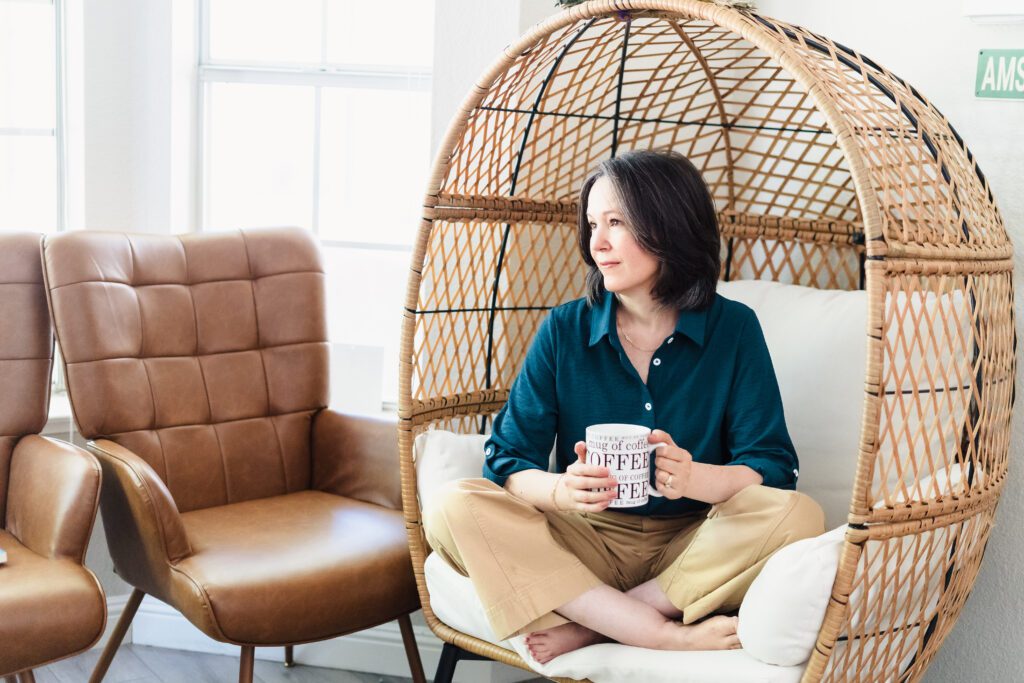 woman sit in a egg chair, holding a cup of coffee and look away through the window