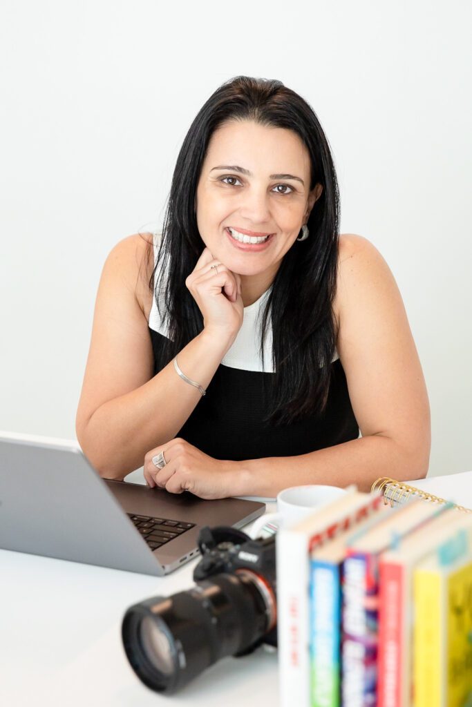 woman sitting in her office, camera, laptop and books on the table, hands in the chin, and smiling